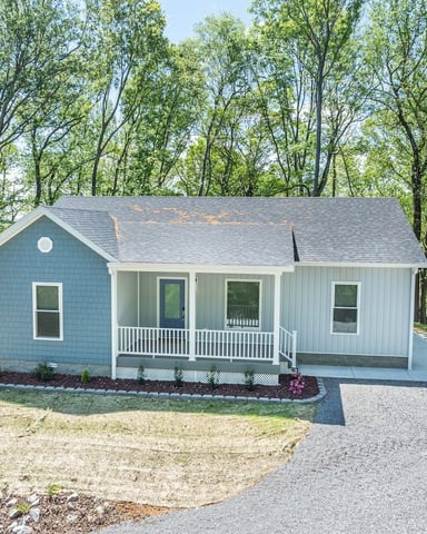 Small blue and white cottage with front porch surrounded by tall trees and gravel driveway