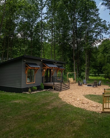 Modern black cabin with wooden porch nestled among tall trees, gravel patio with Adirondack chairs, wooden deck railing in foreground.
