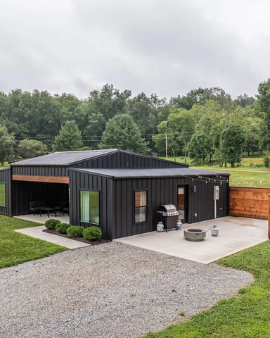 Modern black metal cabin with open-air patio, fire pit, and wooden fence on grassy property surrounded by trees