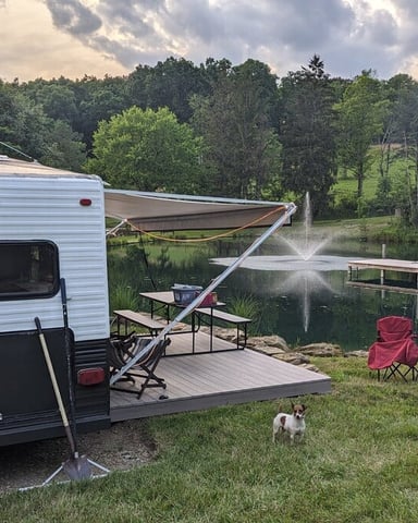 Travel trailer campsite by lake with awning, camping chairs, fire pit, and forested shoreline