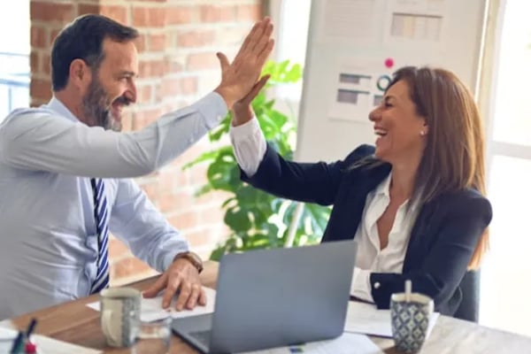 Two professionals high-fiving at a desk with laptop in modern office with brick wall and plant