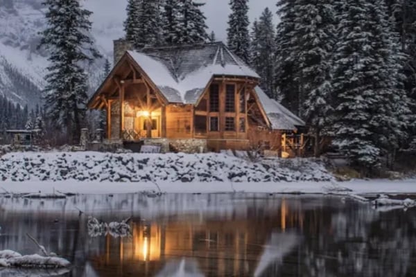 Illuminated wooden cabin with snow-covered roof beside frozen river, surrounded by snow-laden evergreen forest at dusk